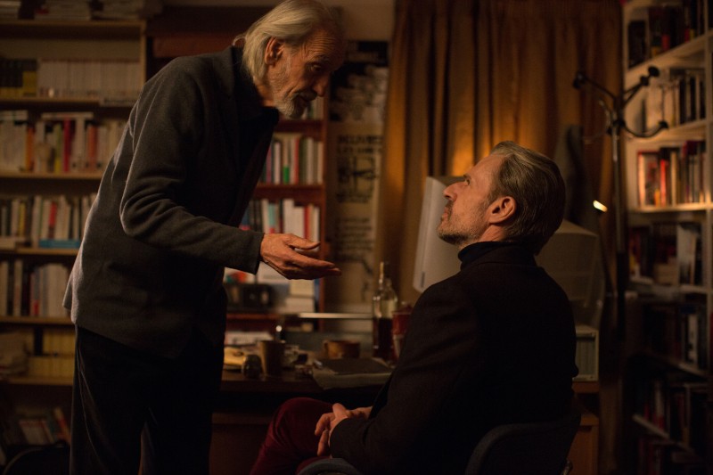 Starszy mężczyzna stoi i gestykuluje, rozmawiając z siedzącym mężczyzną w słabo oświetlonym pomieszczeniu, wypełnionym regałami z książkami i biurkiem, co tworzy poważną atmosferę.