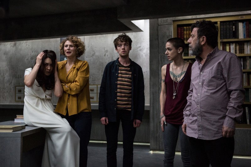 Pięć osób stoi w słabo oświetlonym pomieszczeniu; jedna kobieta ubrana na biało siedzi z głową w dłoniach, podczas gdy pozostali, z poważnymi lub zaniepokojonymi minami, stoją w pobliżu na tle regałów z książkami i szarych ścian.