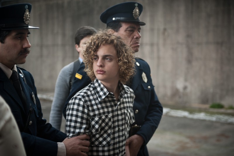 Młody człowiek z kręconymi włosami, ubrany w czarno-białą koszulę w kratkę, jest eskortowany przez umundurowanych policjantów na zewnątrz, z betonowym murem w tle.