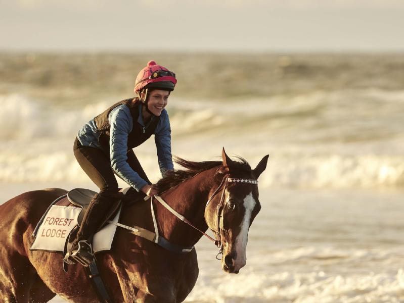 Osoba ubrana w kask i kamizelkę jedzie na koniu z napisem Forest Lodge wzdłuż plaży, w tle widać fale i ocean.
