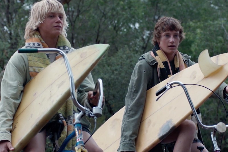 Dwóch młodych ludzi z mokrymi włosami trzyma deski surfingowe i jeździ na rowerach, ręczniki owinięte wokół szyi. Mają na sobie swobodne kurtki i są otoczeni zielonymi drzewami, patrząc w dal.