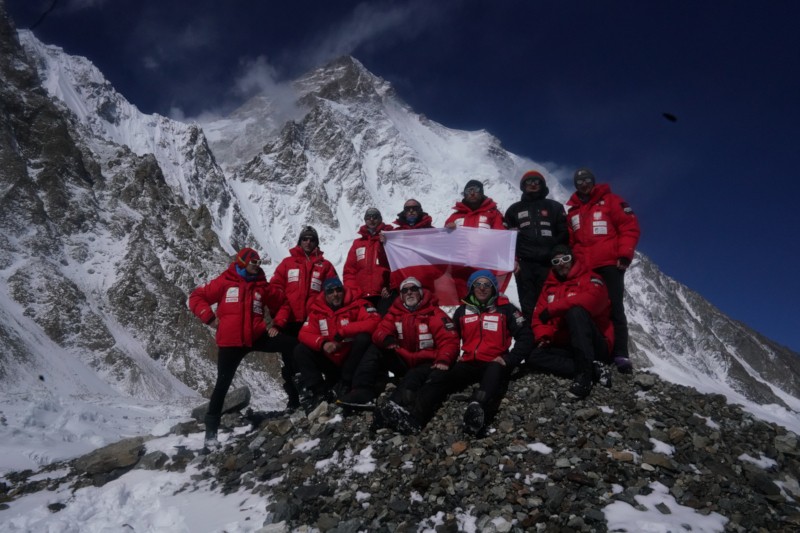 Grupa alpinistów w czerwonych kurtkach pozuje na ośnieżonym, skalistym zboczu na tle pokrytego śniegiem szczytu górskiego, trzymając białą flagę, mając na sobie okulary przeciwsłoneczne i sprzęt zimowy.