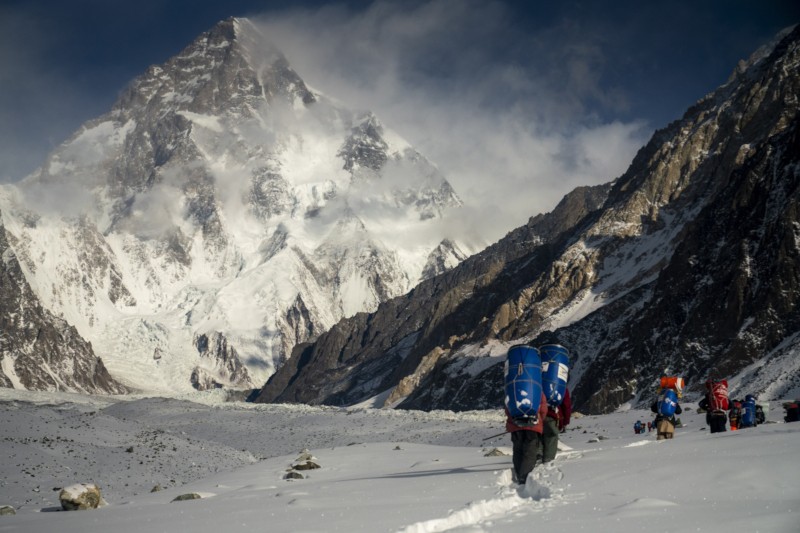 Grupa wspinaczy z plecakami wędruje przez śnieg w kierunku ogromnego, pokrytego śniegiem szczytu górskiego, otoczonego skalistymi grzbietami pod częściowo zachmurzonym niebem.