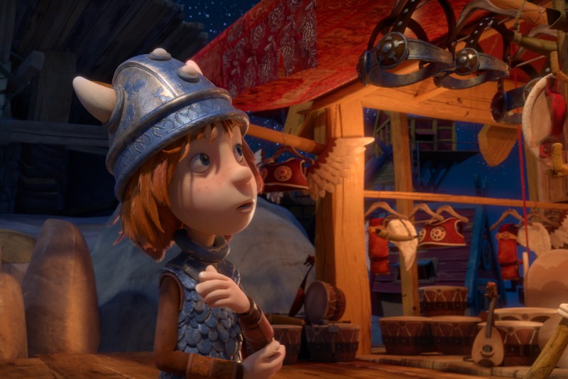 Animowane dziecko w hełmie i zbroi wikinga stoi nocą na kolorowym, ciepło oświetlonym straganie targowym, patrząc w zachwycie na przedmioty wiszące nad nim. W tle widać instrumenty muzyczne i drobiazgi.