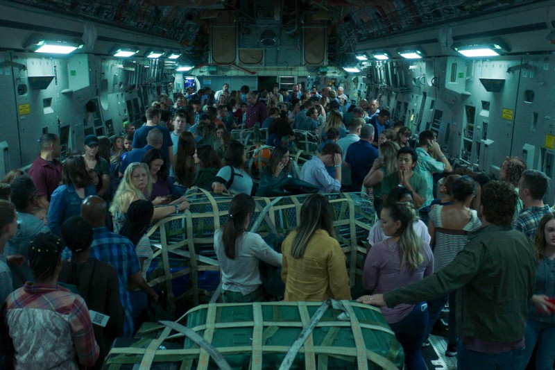 Duża grupa ludzi, składająca się z mężczyzn, kobiet i dzieci, tłoczy się wewnątrz wojskowego samolotu transportowego, w otoczeniu skrzyń i zaopatrzenia, w słabym, niebieskawym świetle.