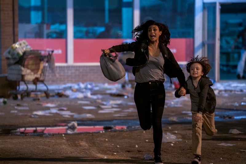 Kobieta i młody chłopak biegną gorączkowo nocą przez zaśmiecony parking, oboje wyglądają na przestraszonych. W tle widać sklep z wybitymi oknami i rozrzuconymi gruzami.