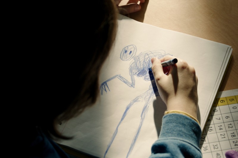 Dziecko rysuje na białym papierze niebieską, abstrakcyjną postać z okrągłą głową i długimi kończynami. Dziecko trzyma niebieską kredkę, a na pobliskim biurku częściowo widoczna jest siatka liczbowa.