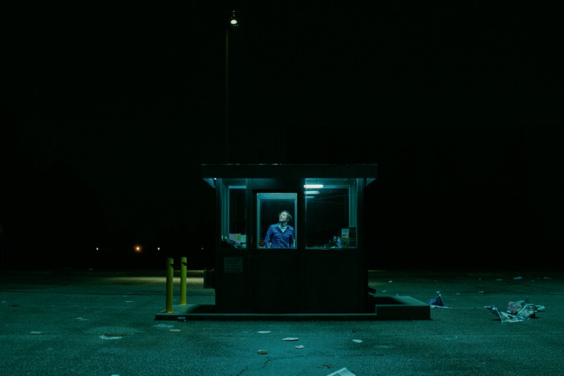 Osoba stoi sama nocą w słabo oświetlonym stoisku na pustym parkingu, otoczona ciemnością i porozrzucanymi śmieciami na ziemi.