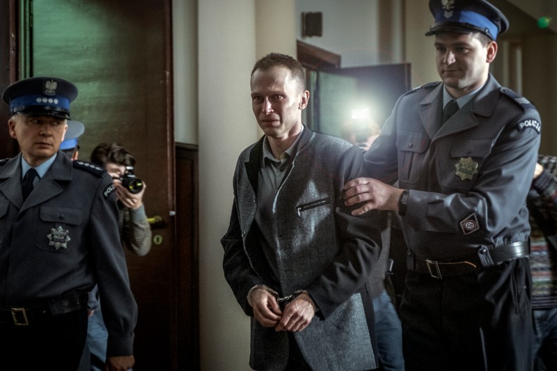 Mężczyzna w kajdankach jest eskortowany przez policjantów przez korytarz, podczas gdy fotografowie uchwycają scenę, a jeden z policjantów trzyma go za ramię. Atmosfera wydaje się napięta i oficjalna.