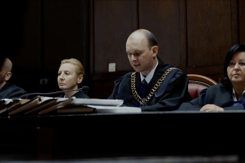 Grupa sędziów w czarnych togach siedzi przy ławie sądowej z dokumentami przed sobą. Sędzia w centrum nosi złoty łańcuch i czyta lub mówi, podczas gdy inni słuchają uważnie. Otoczenie wydaje się formalne i oficjalne.