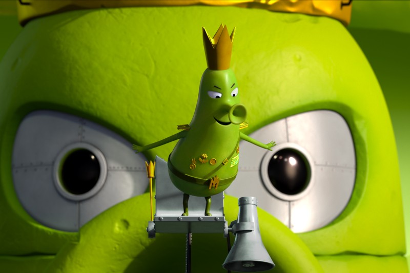 Zielona, kreskówkowa postać w złotej koronie i z medalami pewnie stoi na platformie, w tle widać dużą, surową zieloną twarz z metalowymi powiekami i przenikliwym wzrokiem.