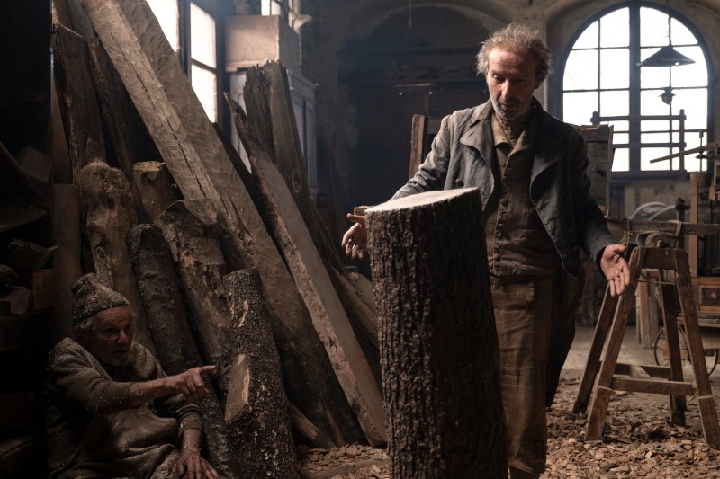 Dwóch mężczyzn w słabo oświetlonym warsztacie otoczonym dużymi drewnianymi deskami; jeden stoi z otwartymi ramionami obok pnia drzewa, podczas gdy drugi siedzi na podłodze, wskazując i patrząc na drewno.