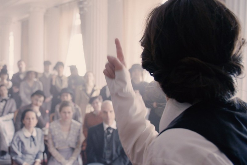 Osoba z długimi włosami i staromodnym ubraniem gestykuluje, przemawiając do siedzącej publiczności w pomieszczeniu z wysokimi kolumnami i miękkim światłem. Publiczność uważnie obserwuje.
