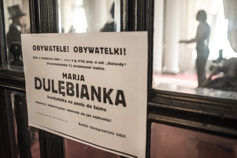 Znak w języku polskim na szklanych drzwiach promuje kandydaturę Marji Dulebianki do Sejmu. Ludzie są widoczni w tle przez szkło, w delikatnie oświetlonym pomieszczeniu.