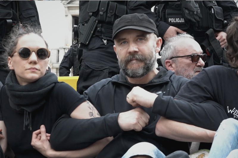 Grupa protestujących siedzi blisko siebie, trzymając się za ręce, zwrócona twarzą do przodu. Za nimi stoją policjanci w czarnych mundurach. Atmosfera wydaje się napięta i poważna.