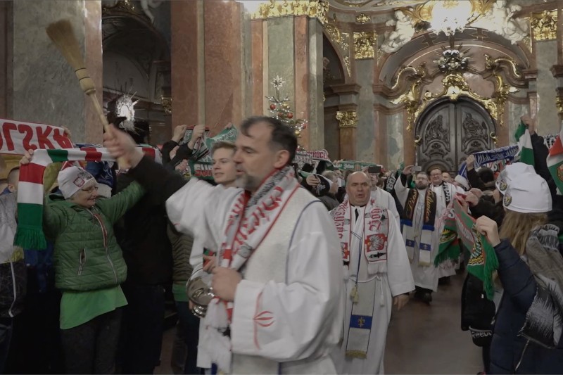 Ksiądz w białych szatach kropi wodą święconą tłum wewnątrz zdobionego kościoła, podczas gdy ludzie ubrani w kolorowe szaliki i czapki podnoszą transparenty i wiwatują. Kościół ma marmurowe kolumny i złote dekoracje.