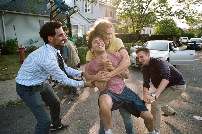 Czterech młodych mężczyzn śmieje się i żartobliwie walczy na podmiejskiej ulicy, a jeden w różowej koszuli jest trzymany przez pozostałych. W tle widać biały samochód z otwartymi drzwiami i dom.