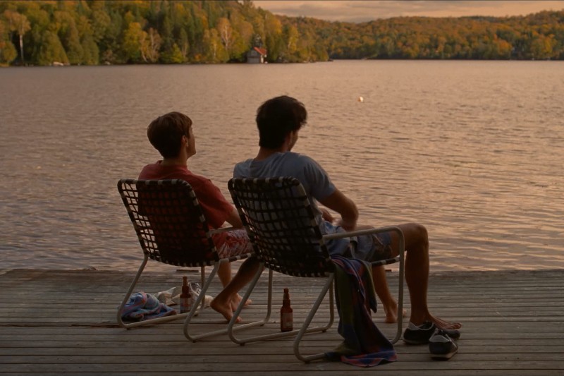Dwie osoby siedzą na krzesłach na drewnianym pomoście, patrząc na spokojne jezioro otoczone drzewami o zachodzie słońca. Ręczniki, buty i butelki z napojami leżą w pobliżu, tworząc spokojną, odprężającą scenę.