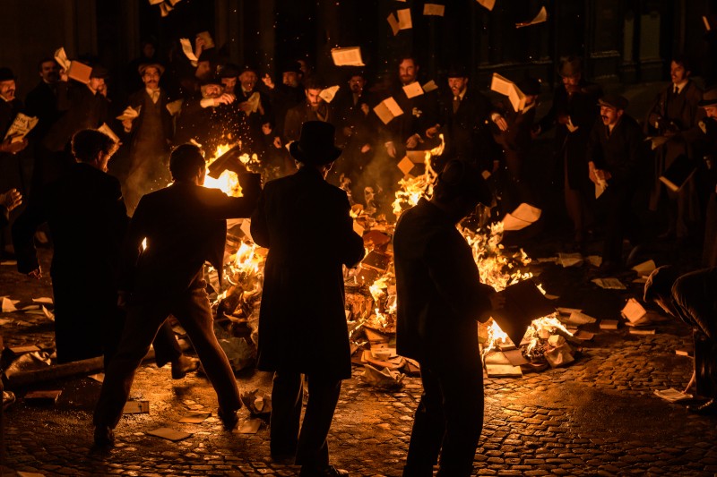 Grupa ludzi w staromodnych ubraniach stoi nocą wokół dużego ognia, paląc papiery i książki, a kolejne strony latają w powietrzu. Płomienie rzucają dramatyczne światło na brukowaną ziemię i otaczające postacie.