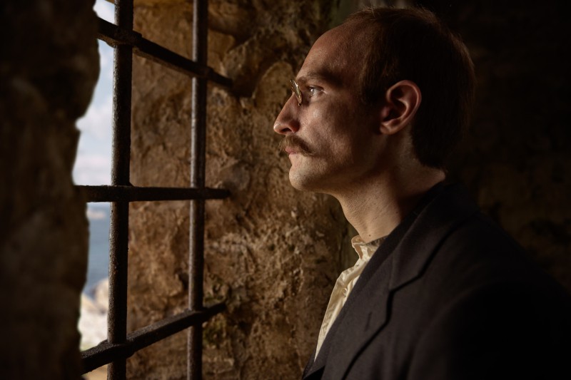 Mężczyzna z wąsami i cofającą się linią włosów stoi z profilu i patrzy przez zakratowane okno z wnętrza kamiennego pokoju. Jego twarz oświetla miękkie światło.