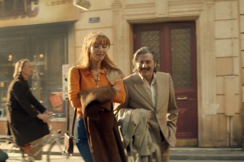 Kobieta z długimi rudymi włosami i mężczyzna w beżowym garniturze idą obok siebie ulicą miasta, oboje trzymając płaszcze. W tle widać rowerzystę i innego pieszego w pobliżu witryny sklepowej.