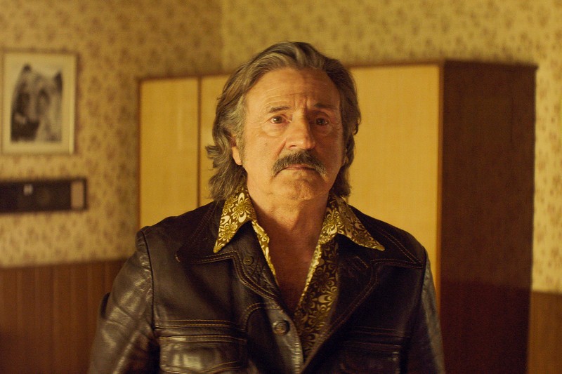 Starszy mężczyzna z siwymi włosami i wąsami, ubrany we wzorzystą koszulę i ciemną skórzaną kurtkę, stoi w pomieszczeniu ze wzorzystą tapetą i drewnianymi meblami. Wygląda na lekko zaniepokojonego.