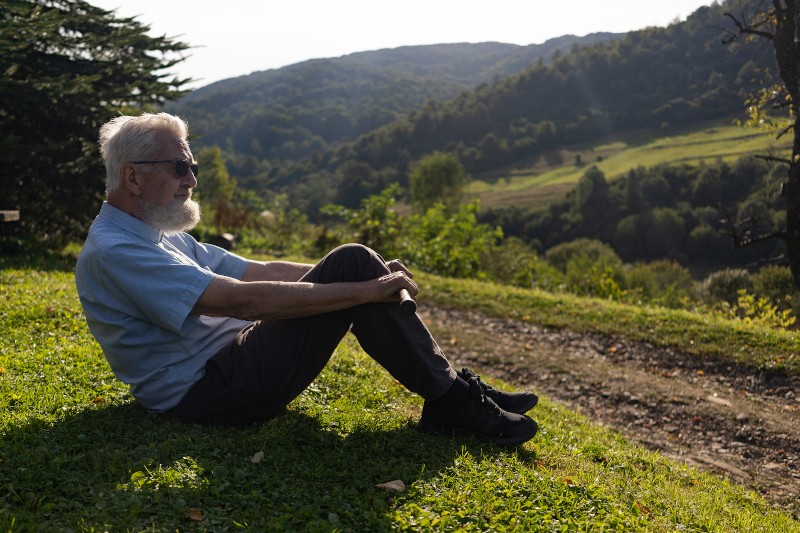 Starszy mężczyzna z siwymi włosami i brodą siedzi na trawie, w zamyśleniu wpatrując się w malowniczy krajobraz zielonych wzgórz pod czystym niebem. Światło słoneczne rzuca miękkie cienie, tworząc spokojny, kontemplacyjny nastrój.