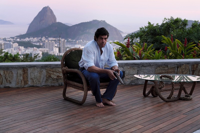 Mężczyzna w białej koszuli i dżinsach siedzi boso na wiklinowym krześle, trzymając pistolet, a obok niego stoi szklany stolik. Scena ukazuje widok na Rio de Janeiro, a w tle widać Górę Sugarloaf o zachodzie słońca.