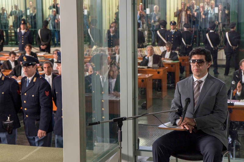 Mężczyzna w garniturze i okularach przeciwsłonecznych siedzi w szklanej obudowie z mikrofonem, naprzeciwko sali sądowej pełnej ludzi, policjantów i reporterów znajdujących się za szklanymi ścianami.