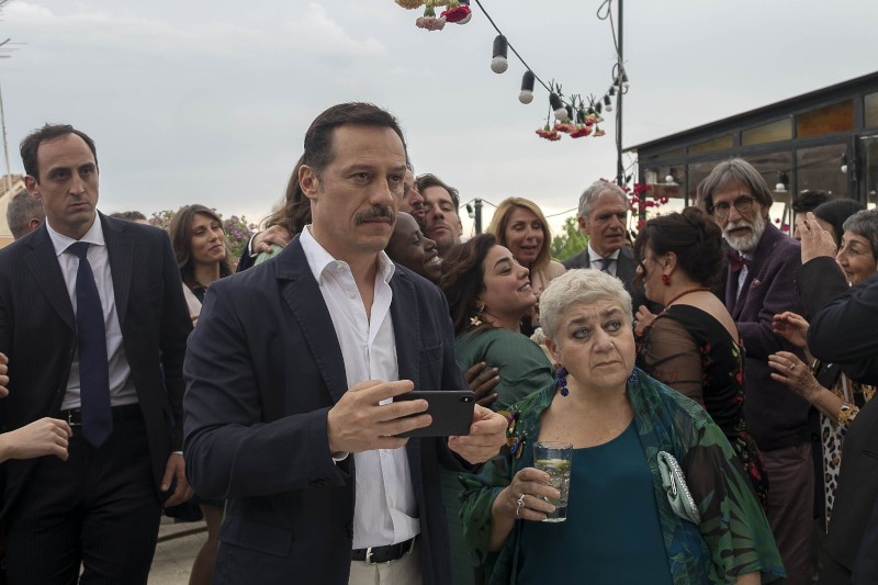 Mężczyzna w garniturze trzymający telefon i starsza kobieta z krótkimi siwymi włosami trzymająca stoisko z napojami pośród tłumu ludzi na imprezie plenerowej, nad głowami wiszą sznury świateł i kwiaty.