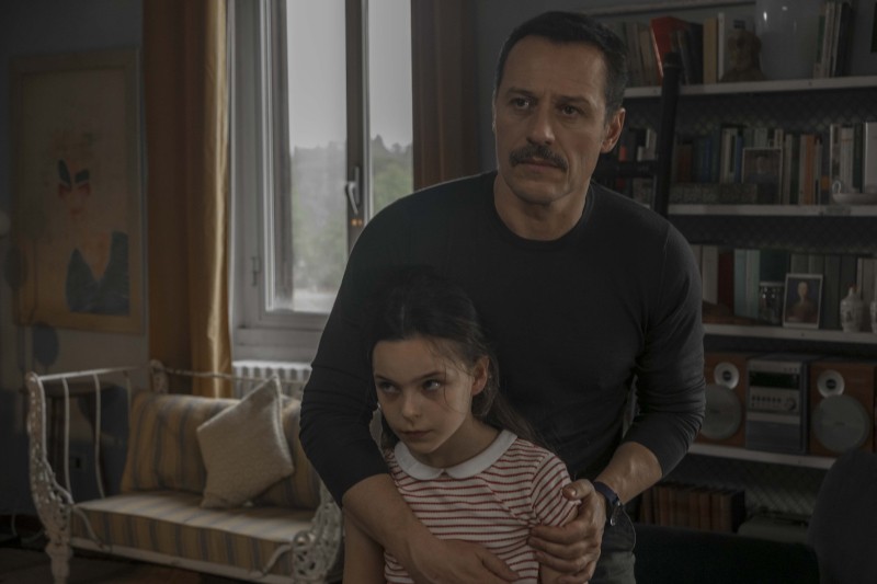 Mężczyzna z wąsami trzyma opiekuńczo młodą dziewczynę w słabo oświetlonym salonie, oboje wyglądają na zaniepokojonych. W tle widać regały na książki, kanapę i okno.