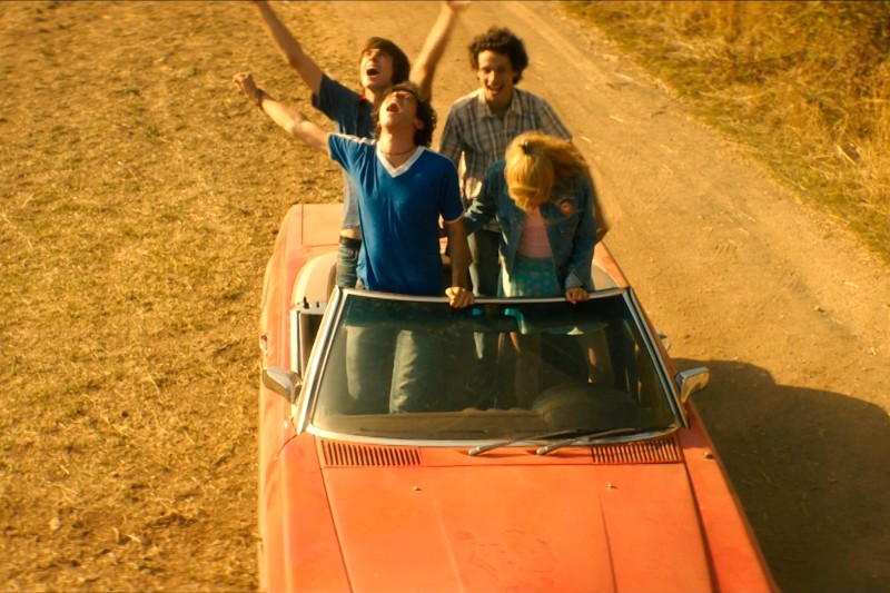 Czwórka młodych ludzi radośnie jedzie czerwonym kabrioletem po drodze gruntowej, w tym dwie osoby stojące i wiwatujące przez otwarty dach. To uwiecznia beztroską, pełną przygód chwilę w jasnym świetle słonecznym.