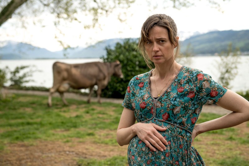 Kobieta w kwiecistej sukience stoi na zewnątrz, trzymając się za ciążowy brzuch i wyglądając na zaniepokojoną. W tle krowa pasie się w pobliżu jeziora, a w oddali widać góry.