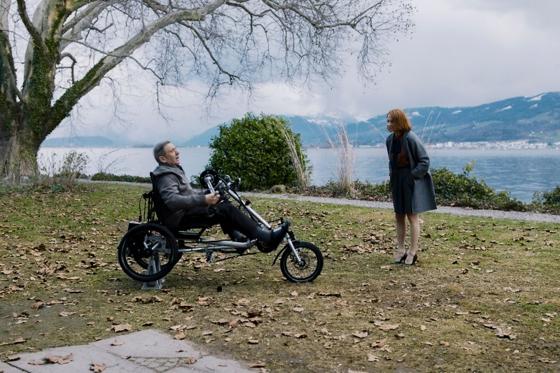 Mężczyzna na trójkołowym wózku inwalidzkim z napędem elektrycznym stoi naprzeciwko kobiety stojącej na pokrytym liśćmi trawniku w pobliżu jeziora, w otoczeniu nagich drzew i w oddali wzgórz pod zachmurzonym niebem.