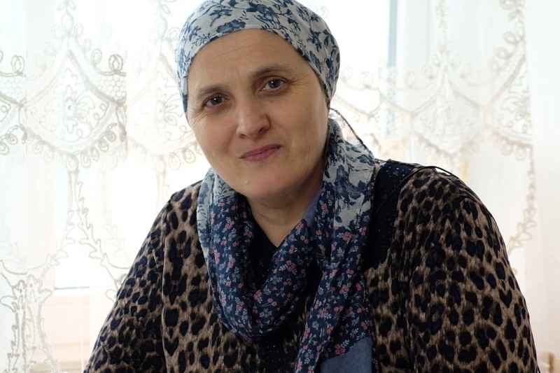 Kobieta ubrana we wzorzystą chustę na głowie i bluzkę z nadrukiem w panterkę siedzi przed koronkową zasłoną i delikatnie uśmiecha się do aparatu.