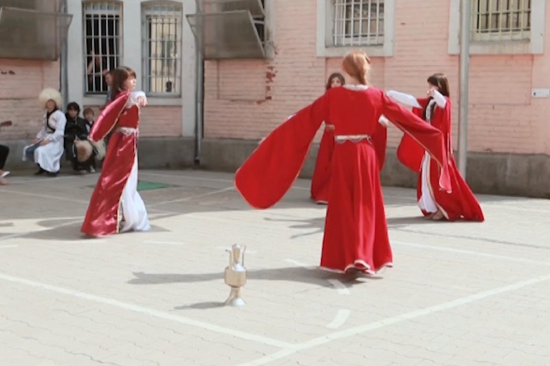 Cztery kobiety w zwiewnych czerwonych sukienkach wykonują tradycyjny taniec na zewnątrz na wybrukowanym terenie, ze srebrnym dzbanem położonym na ziemi w centrum. Kilka osób obserwuje z tła, w pobliżu ceglanego budynku.