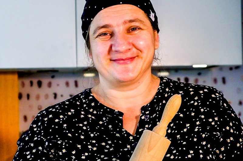 Kobieta ubrana w czarno-białą wzorzystą koszulę i czarną chustę na głowie uśmiecha się, trzymając drewniany wałek do ciasta w kuchni.