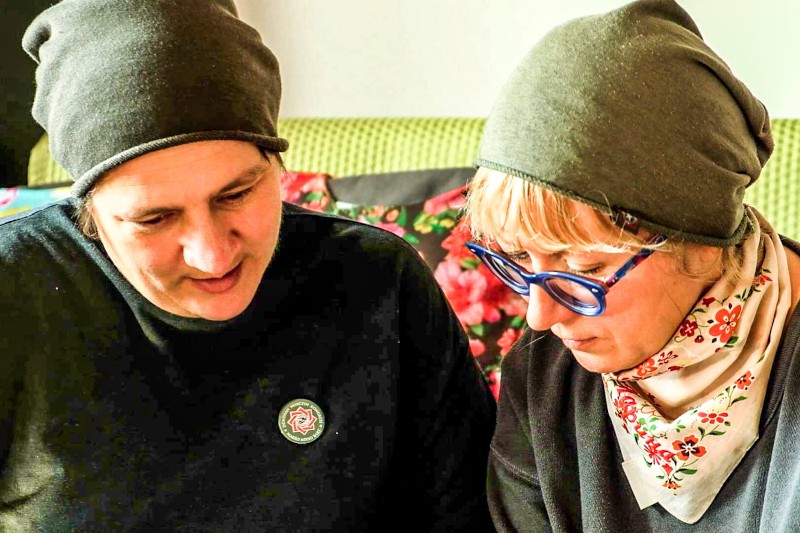 Dwie osoby w ciemnych kapeluszach i swetrach siedzą blisko siebie, patrząc w dół. Jedna ma niebieskie okulary i kwiatowy szalik. Wyglądają, jakby były skupione lub czytały coś poza ramką, a za nimi leży kwiatowa poduszka.