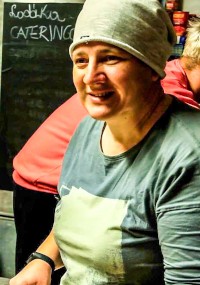 Osoba ubrana w szarą czapkę i koszulkę z krótkim rękawem uśmiecha się w kuchni, w tle widać tablicę z napisem CATERING.