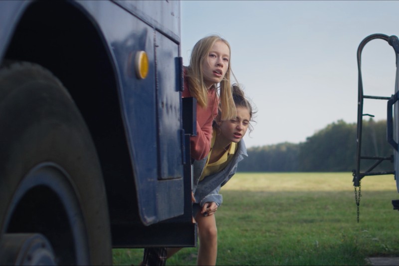 Dwie dziewczyny wychyla się z drzwi niebieskiego autobusu, uważnie patrząc na coś w oddali. Stoją na otwartym trawiastym polu, z drzewami i czystym niebem w tle.