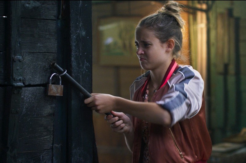 Młoda dziewczyna z blond włosami spiętymi w kok krzywi się, próbując rozbić kłódkę na drewnianych drzwiach łomem w słabo oświetlonym pokoju.