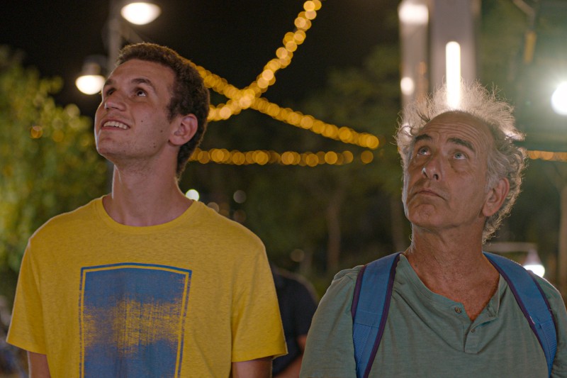 Dwóch mężczyzn stojących na zewnątrz nocą, patrzących w górę. Młodszy mężczyzna po lewej ma na sobie żółtą koszulkę, podczas gdy starszy mężczyzna po prawej ma siwiejące włosy, niebieską koszulę i plecak. W tle świecą lampki.