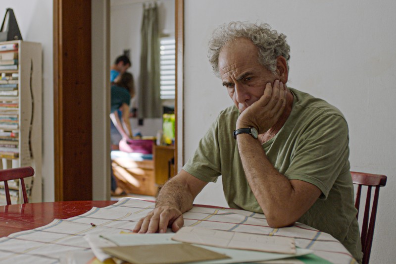 Starszy mężczyzna siedzi przy stole, wygląda na zamyślonego i opiera głowę na dłoni, przed nim leżą papiery. W tle dwie osoby stoją przy łóżku w innym pokoju.