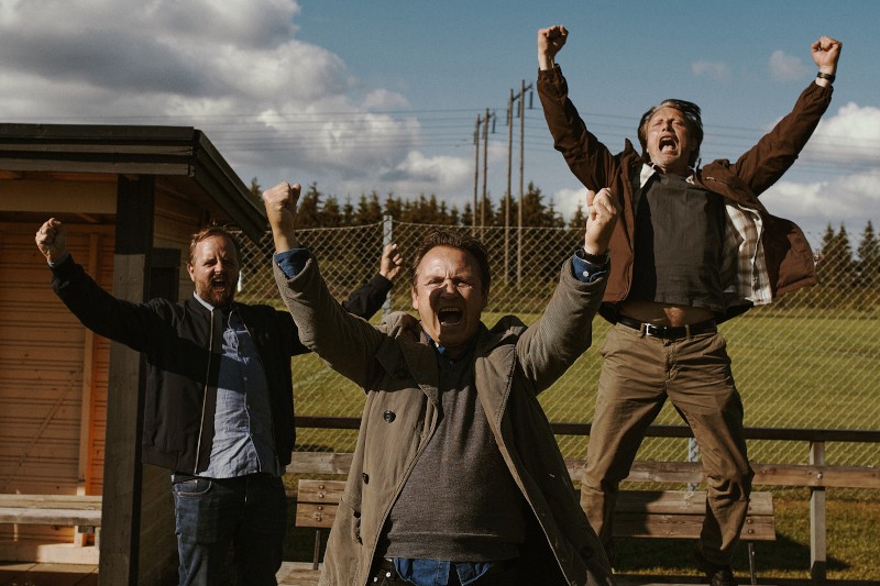 Trzech mężczyzn na zewnątrz świętuje z entuzjazmem, podnosząc ramiona i wiwatując. Jeden mężczyzna podskakuje z podniecenia. Stoją przy ławce i drucianym płocie z zieloną trawą i drzewami w tle.