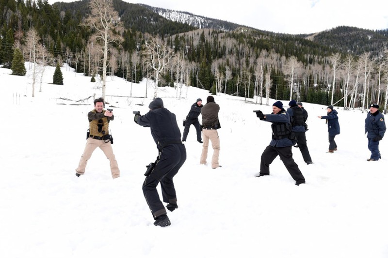 Kilka osób w strojach taktycznych stoi na zewnątrz, na zaśnieżonym terenie, celując do siebie z broni krótkiej w ramach ćwiczeń, na tle drzew i gór.