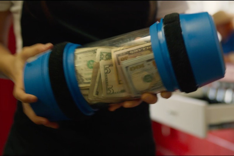 Osoba trzyma niebieską plastikową tubę na gotówkę wypełnioną banknotami dolarowymi o różnych nominałach, często używaną do bezpiecznego transportu pieniędzy w sklepach detalicznych i bankach.