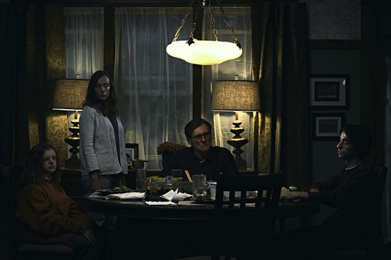 Cztery osoby siedzą przy słabo oświetlonym stole, wyglądając na napiętych i poważnych. Na stole leży jedzenie i naczynia, a nad nimi wisi duża lampa, rzucająca nastrojowe światło. Ciężkie zasłony i lampy wypełniają tło.