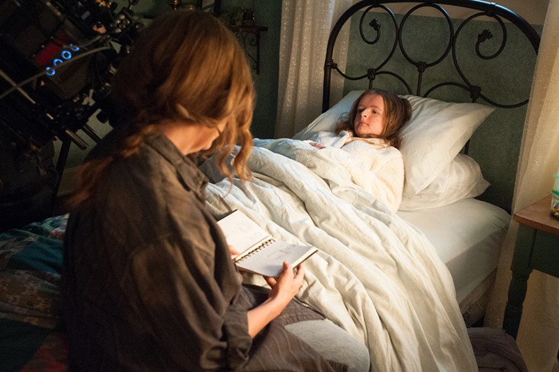 Kobieta siedzi przy łóżku, trzymając notatnik, podczas gdy młoda dziewczyna w białym szlafroku leży pod kołdrą i patrzy na nią. Widoczny jest sprzęt do filmowania, co wskazuje na plan filmowy.