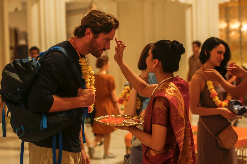 Kobieta w tradycyjnym stroju nakłada tilak na czoło mężczyzny podczas ceremonii powitalnej. Mężczyzna nosi plecak i girlandę kwiatów. W tle widać inne osoby i ciepłe oświetlenie.