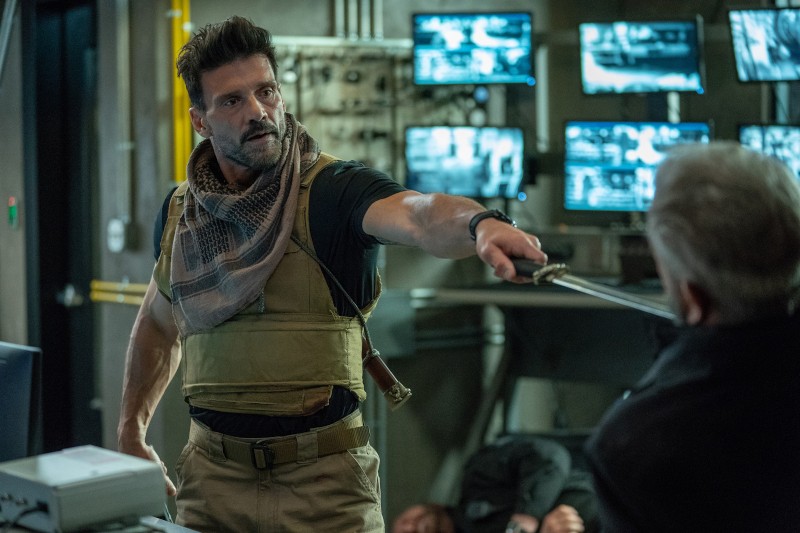 Mężczyzna ubrany w sprzęt taktyczny mierzy mieczem do innego mężczyzny siedzącego w słabo oświetlonym pomieszczeniu z monitorami na ścianach; w tle inna osoba leży nieprzytomna na podłodze.